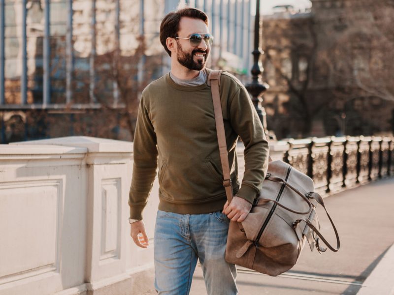 handsome stylish hipster man walking in city street with leather bag wearing sweatshot and sunglasses, urban style trend, sunny day, smiling happy traveler